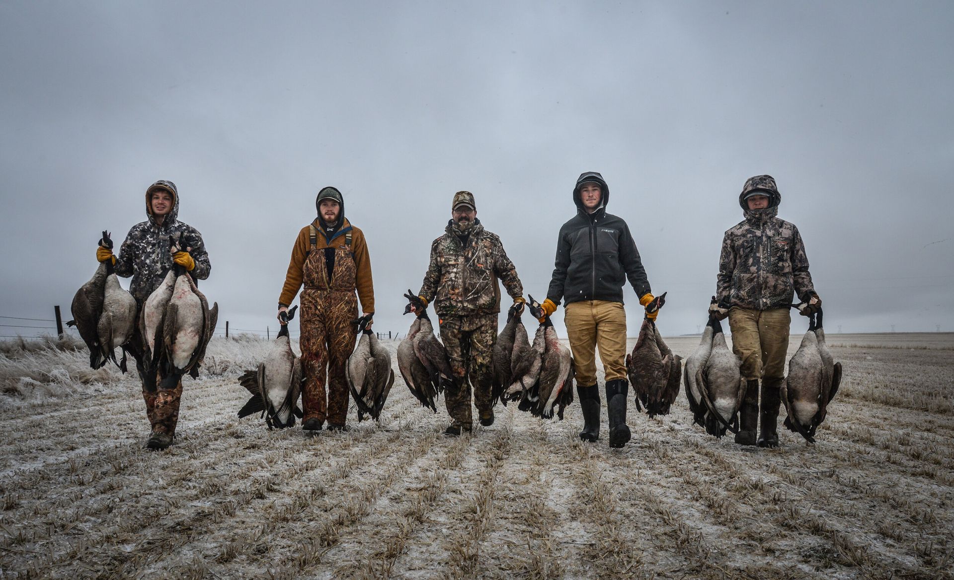 Six people walking toward camera across a muddy field under a gray sky, carrying a deer
