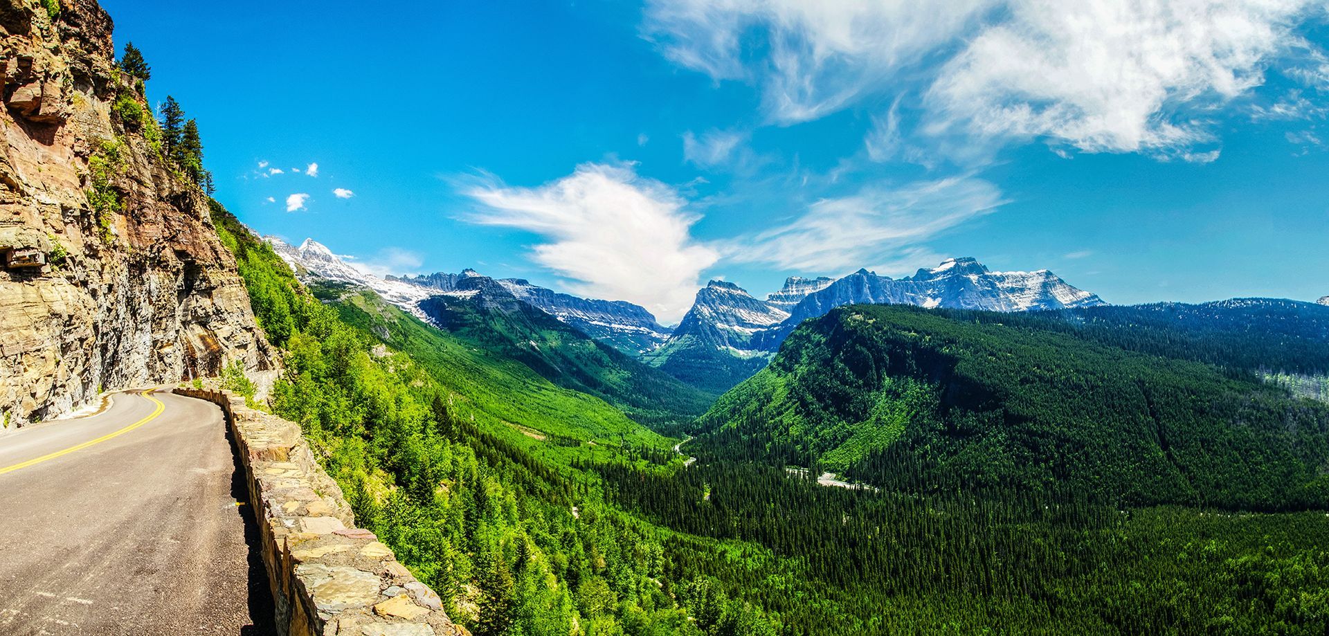 Mountain road overlooking a green valley and snow-capped peaks under a blue sky