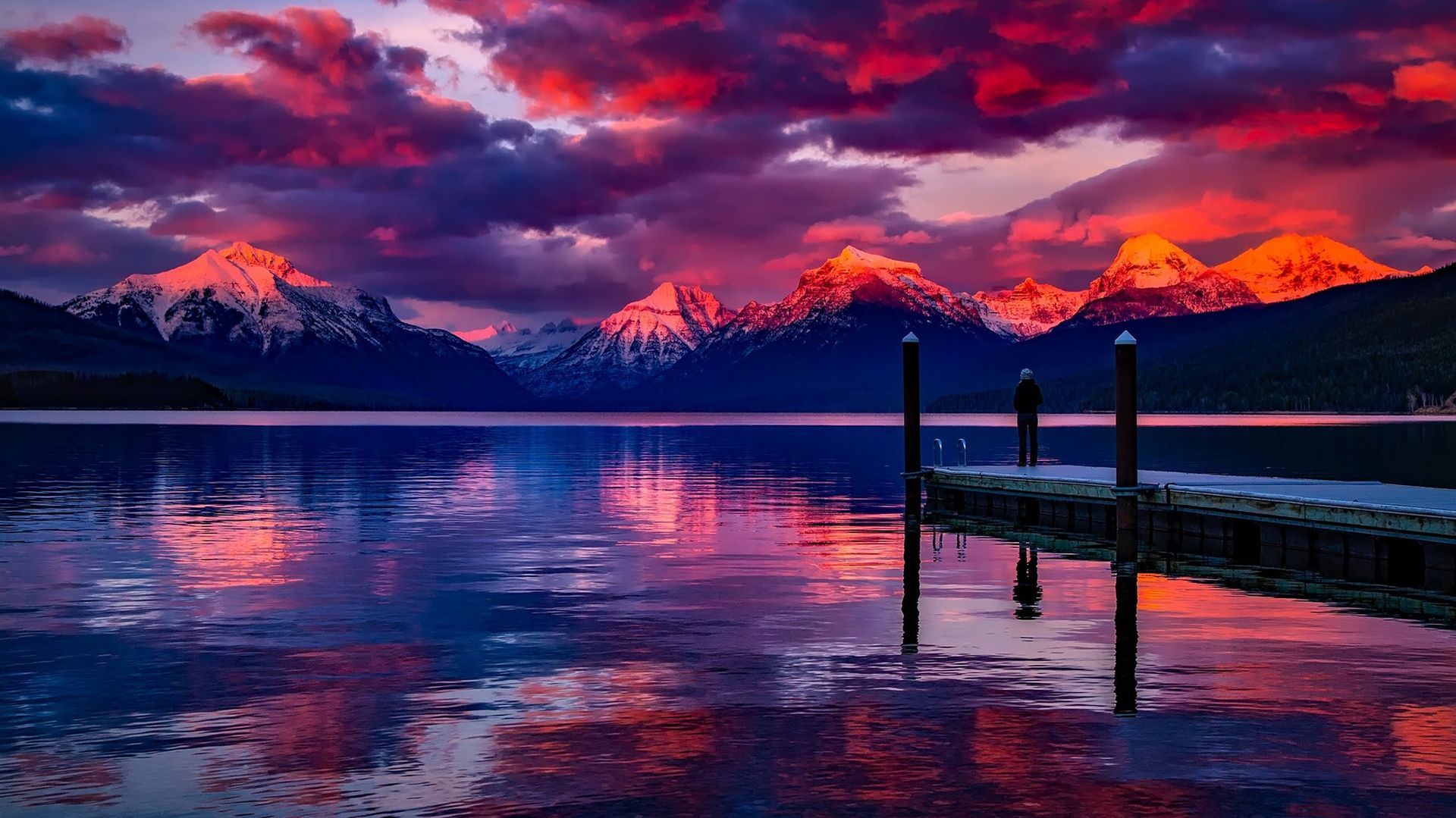 Pink sunset over snowy mountains reflected in a calm lake, with a dock in the foreground.
