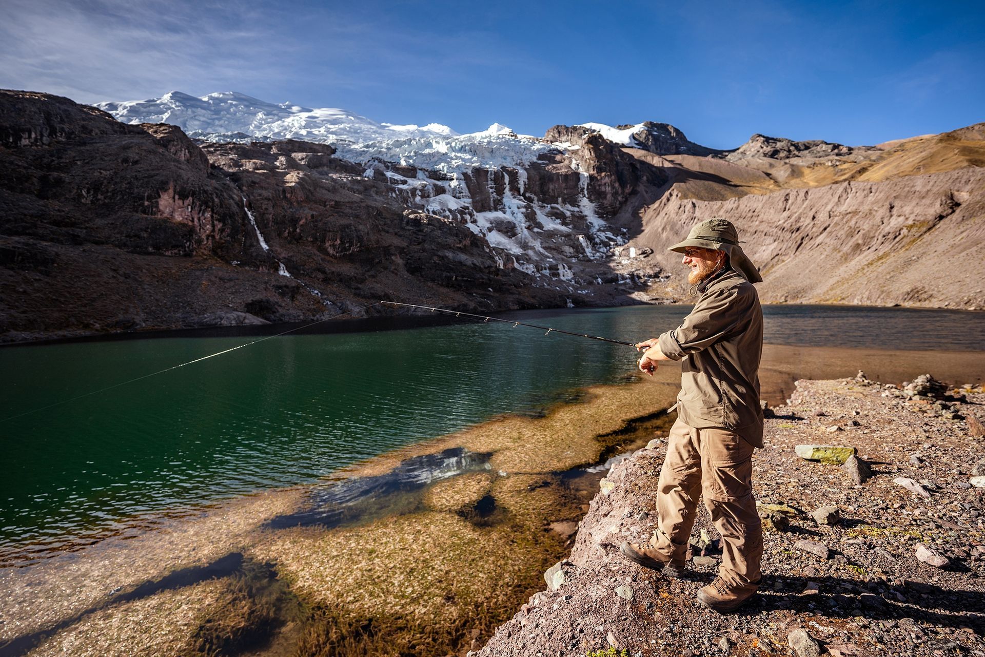 Person fishing beside a turquoise mountain lake with snow-covered peaks in the background
