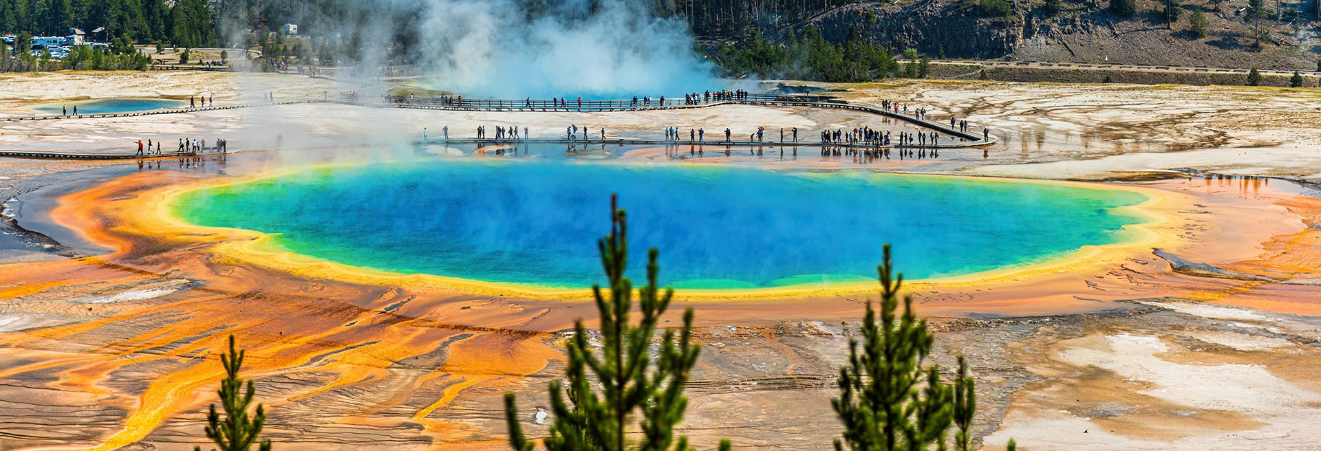 Colorful hot spring with turquoise center, orange mineral edges, steam rising, and trees in the foreground