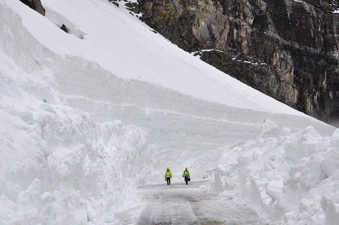 Two hikers in yellow jackets walking through a snowy mountain pass beneath an icy slope