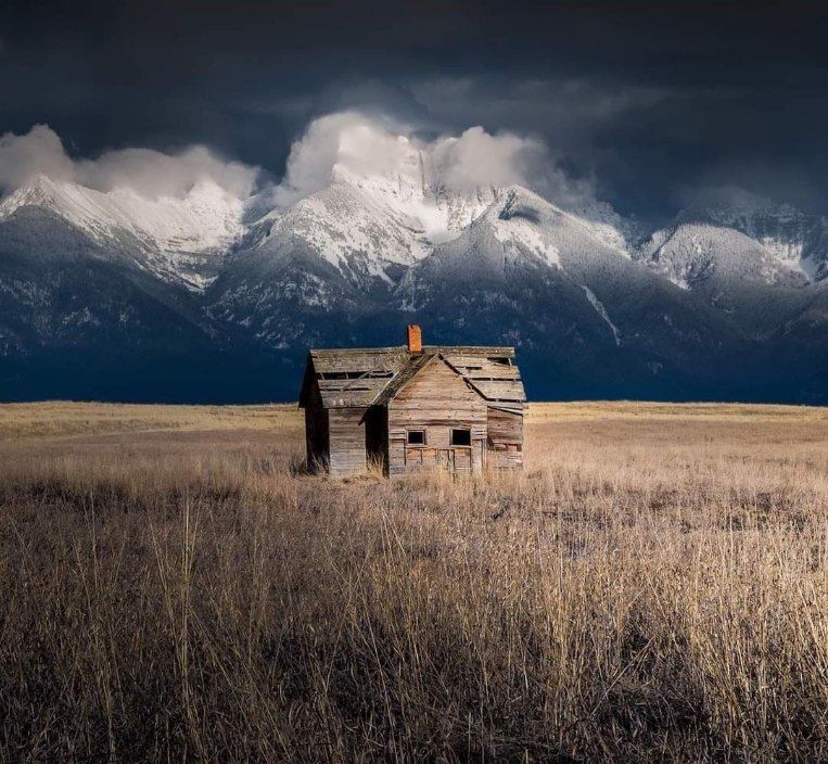 Small weathered cabin in a grassy field with snowy mountains and dark clouds in the background