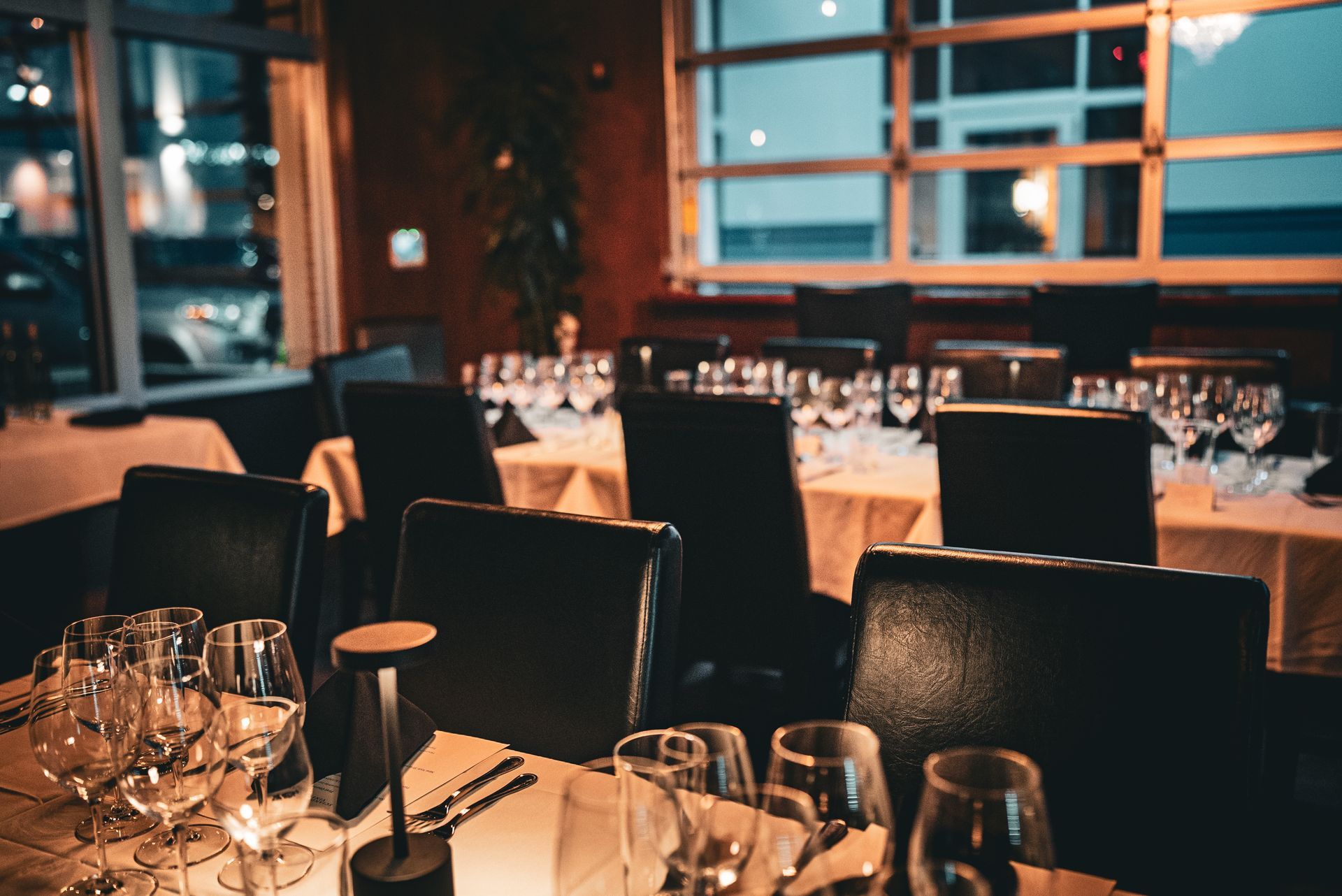 Elegant restaurant interior with set tables, chairs, and glasses, near a window. Dark decor, warm lighting.