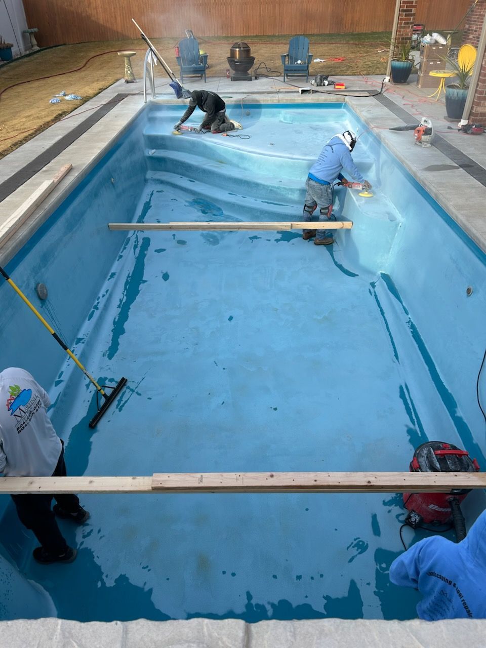 Workers renovating a blue swimming pool, using tools and equipment, in an outdoor setting.