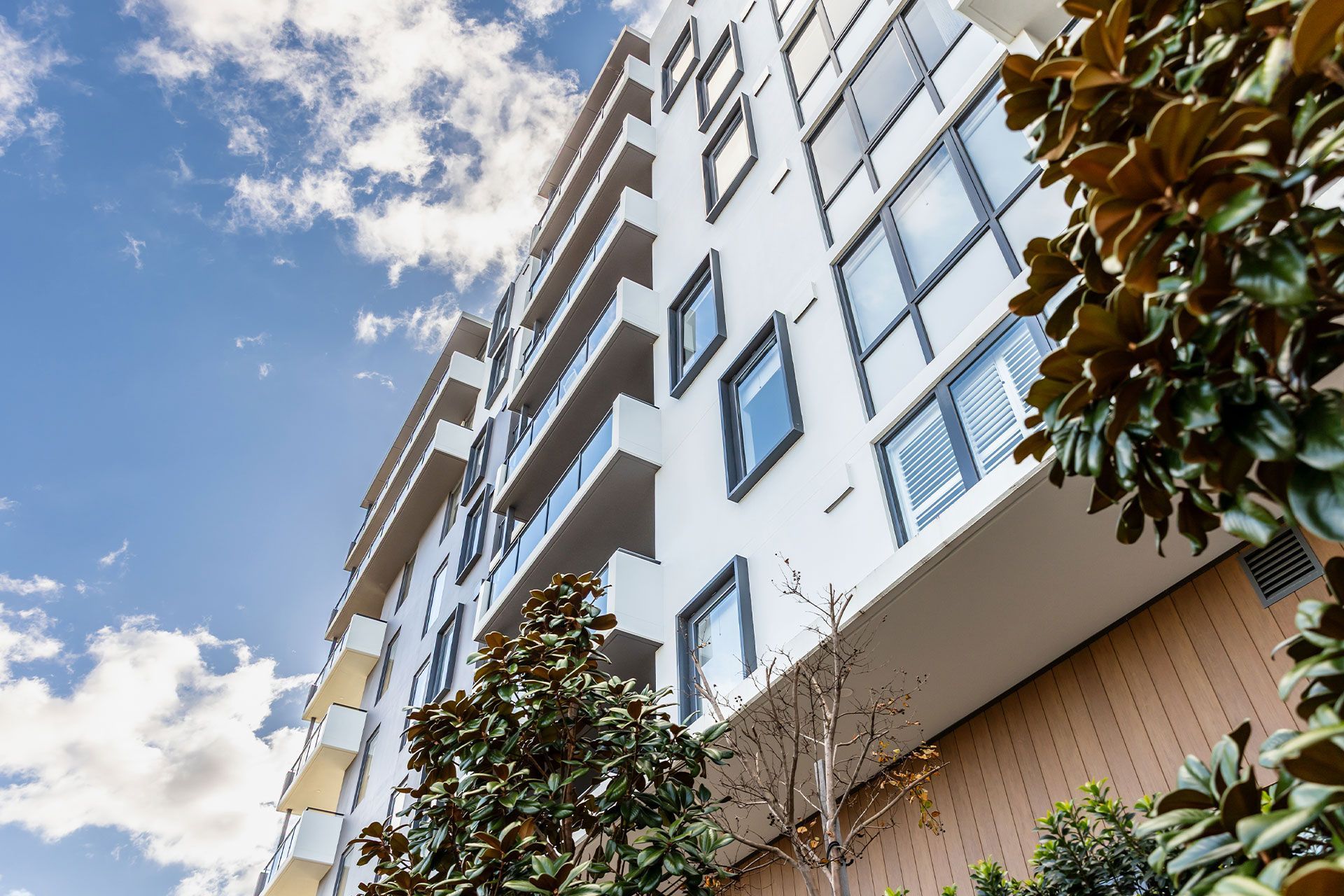 Modern white apartment building with balconies, blue sky, and foliage.