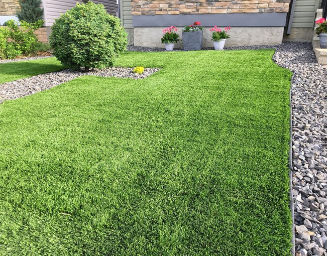 Lush green artificial grass lawn with decorative gravel borders and foundation of a house.