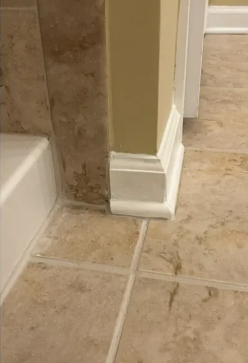 Bathroom corner with beige tile floor, off-white baseboard, and tan wall.