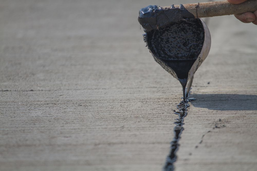 A person fills a crack in concrete with black sealant using a trowel.