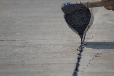 A person fills a crack in concrete with black sealant using a trowel.