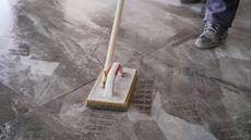 Person cleaning a marble tile floor with a scrub brush and handle.