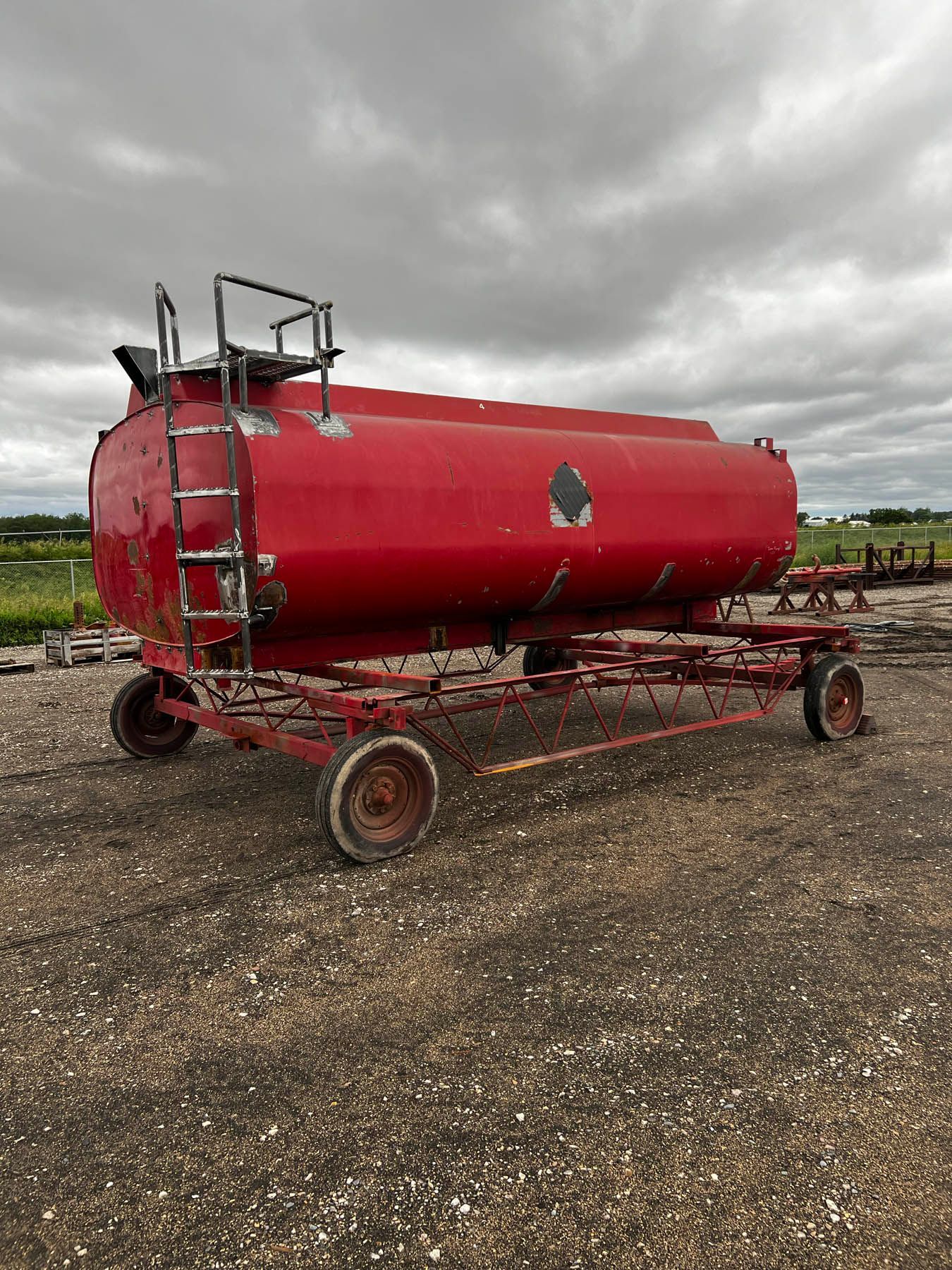 Red tank on wheels with a ladder, sitting outdoors on gravel under a cloudy sky.