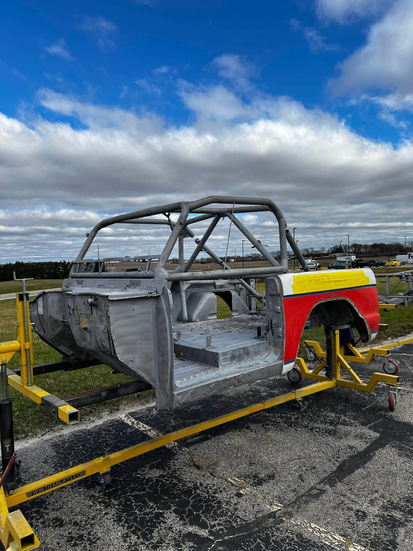 A silver car frame with a roll cage sits on a yellow stand outside under a cloudy sky. The rear quarter panel is red, yellow, and white.