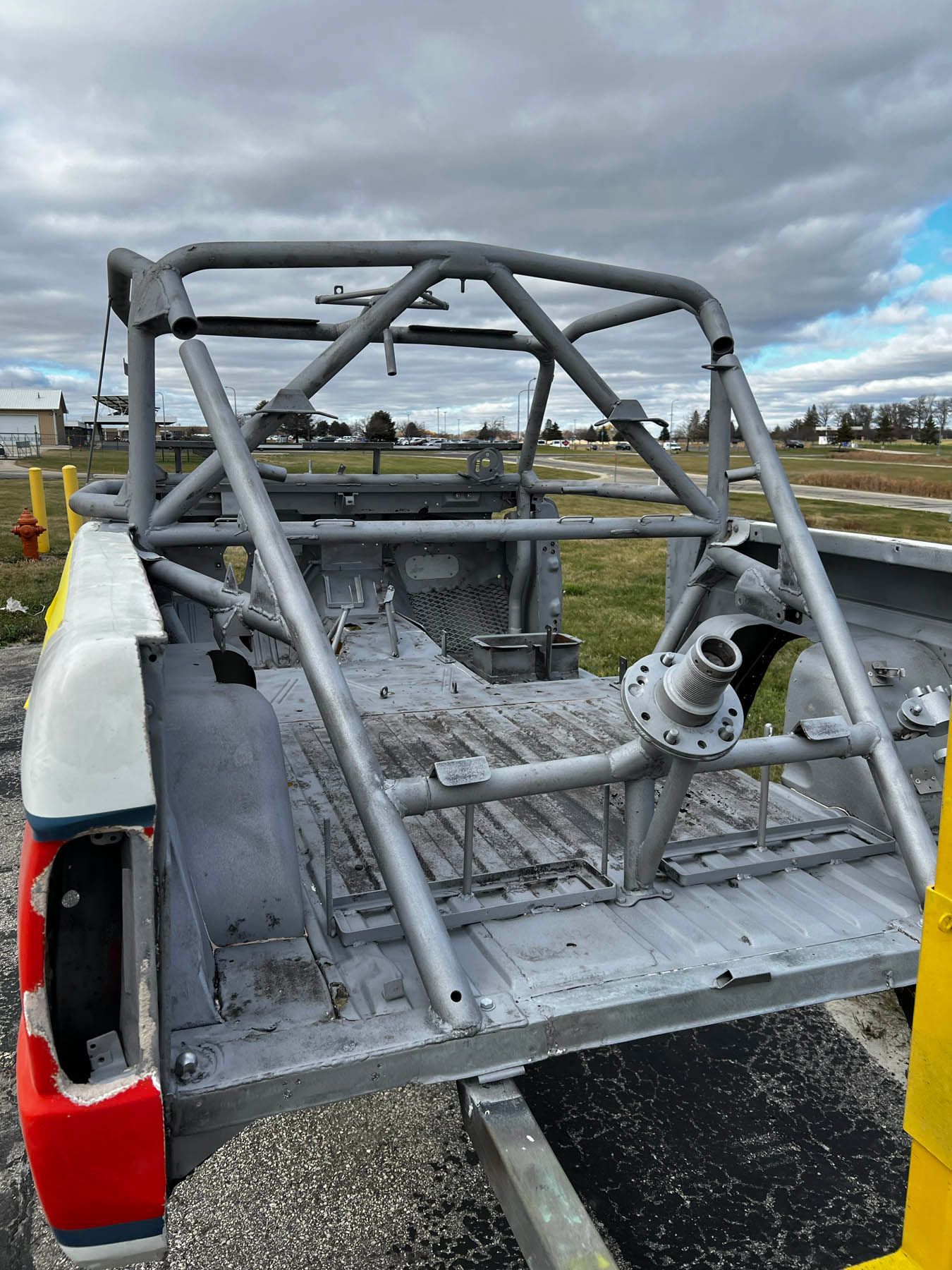 Gray modified pickup truck bed with a roll cage on a cloudy day. The bed is unfinished with a rear axle visible.
