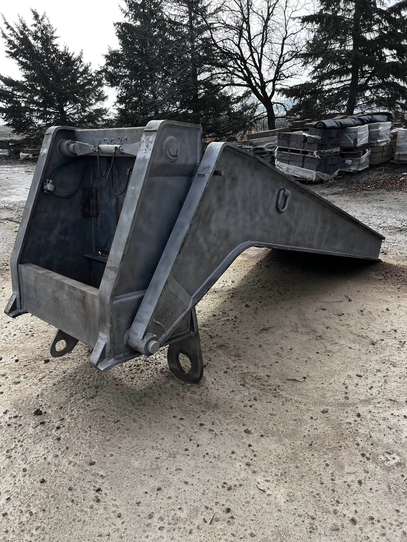Large, gray metal bucket, likely for heavy machinery, resting on a gravel surface outdoors.