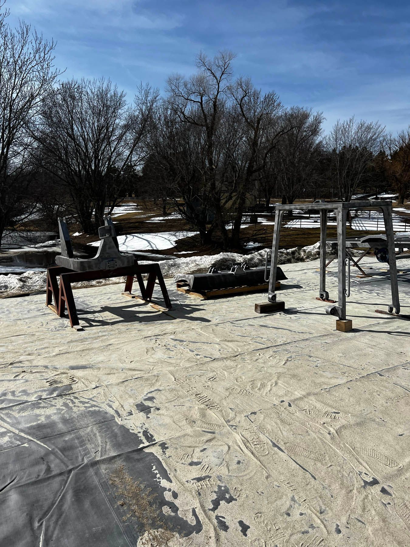 Outdoor scene with various metal structures and tools set up on a light-colored surface, partially covered by a dark material, in a snowy, barren landscape.