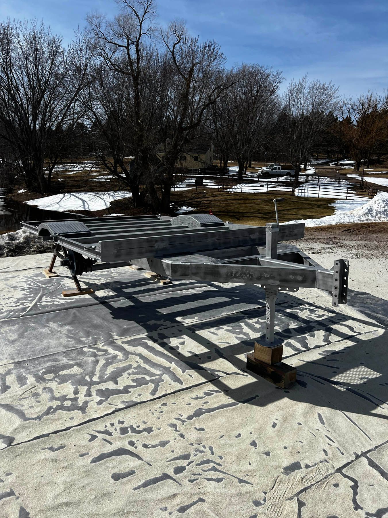 Gray utility trailer parked on a concrete surface, set in a snowy field with bare trees in the background under a blue sky.