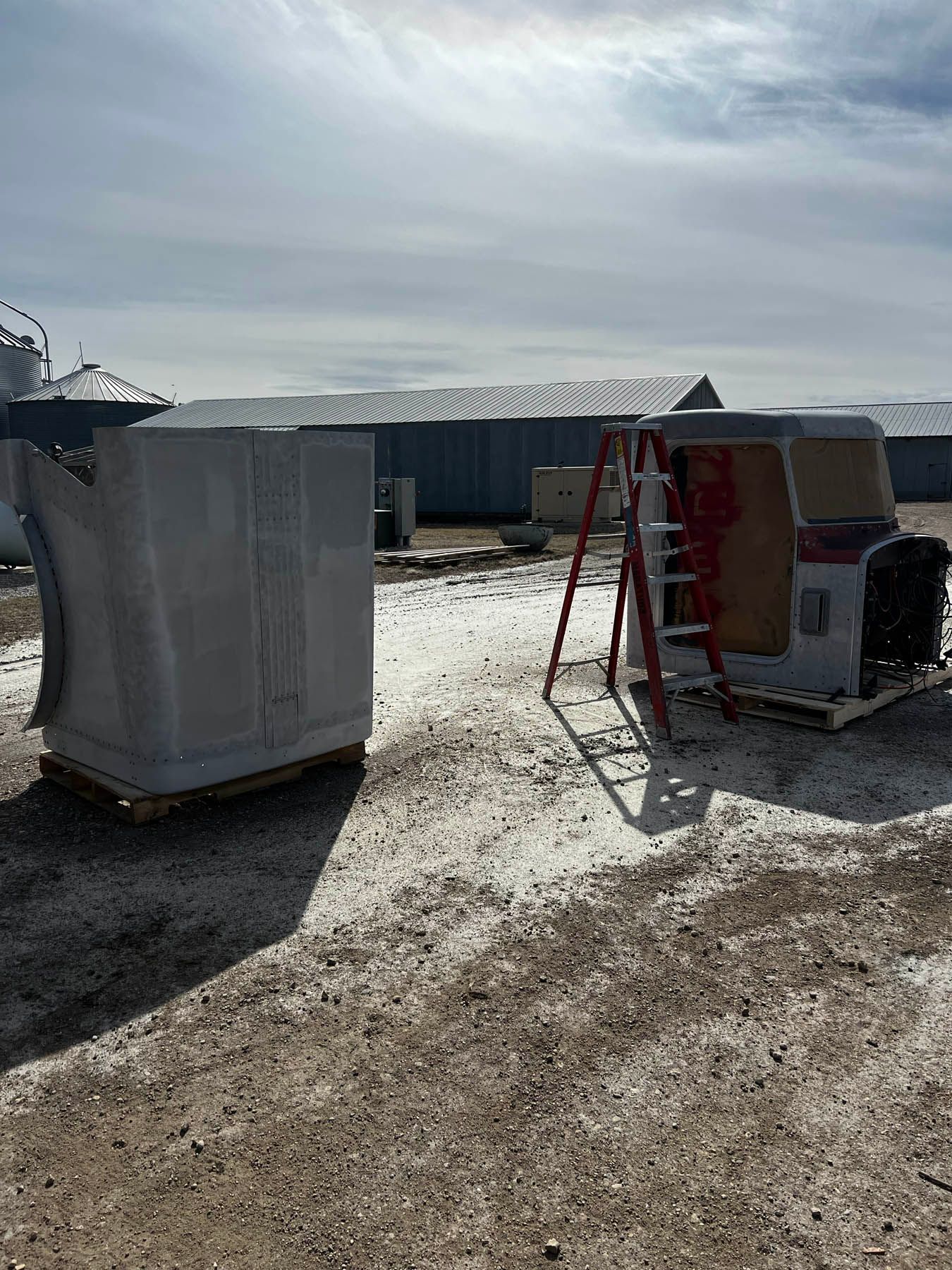 Two large truck parts on pallets, a ladder, and buildings in a sunny outdoor setting. One part has red interior.