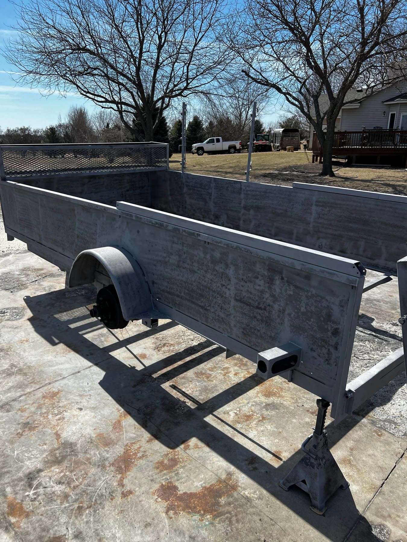 Gray utility trailer outdoors, supported by jack stands. The trailer bed is visible, as is the trailer hitch.