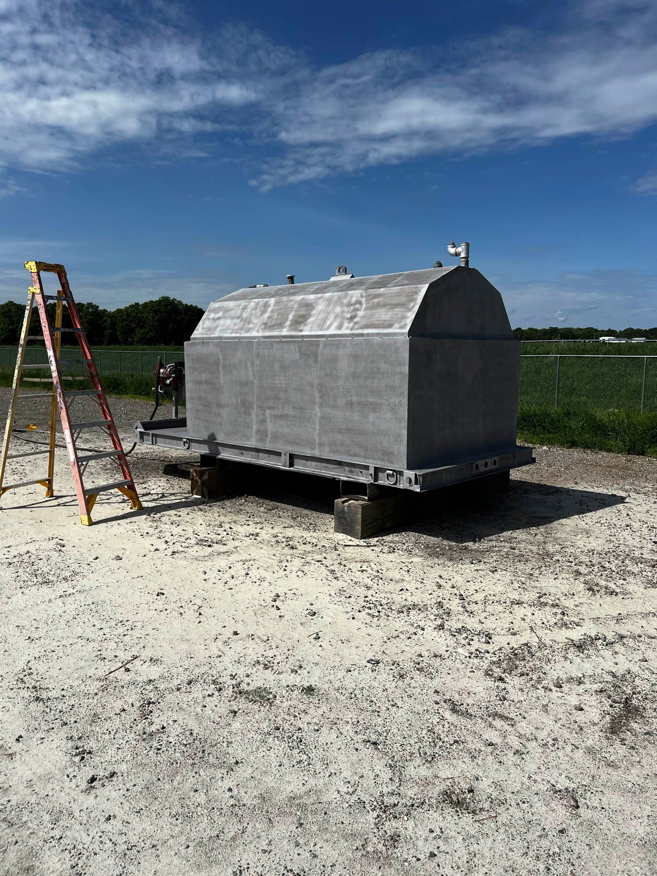 Gray concrete structure on a trailer with a ladder beside it, set on a gravel area under a blue sky.