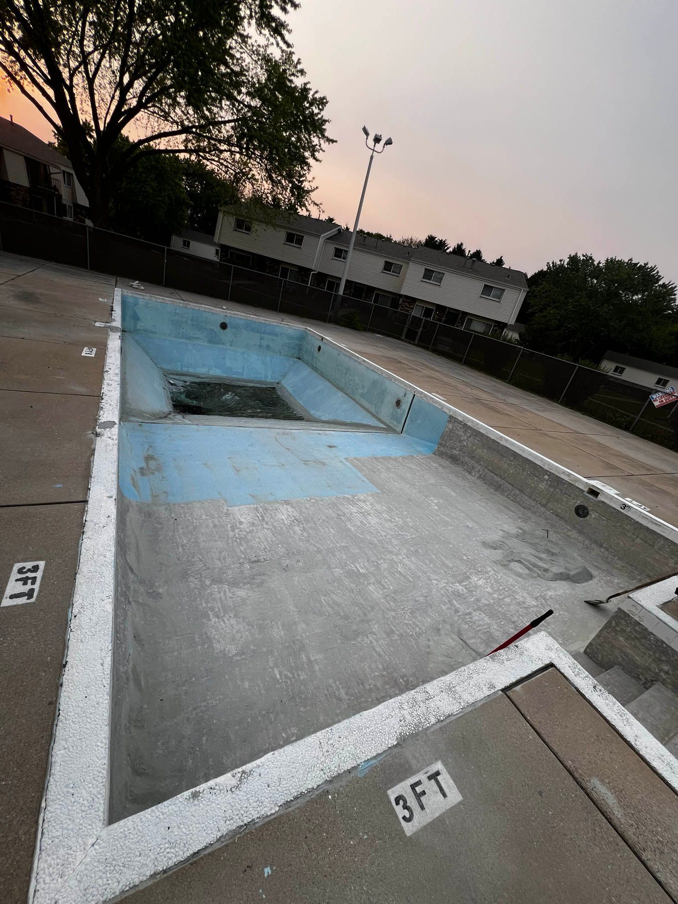 Empty, concrete swimming pool with faded blue paint. Sun sets in the background over low buildings and trees.