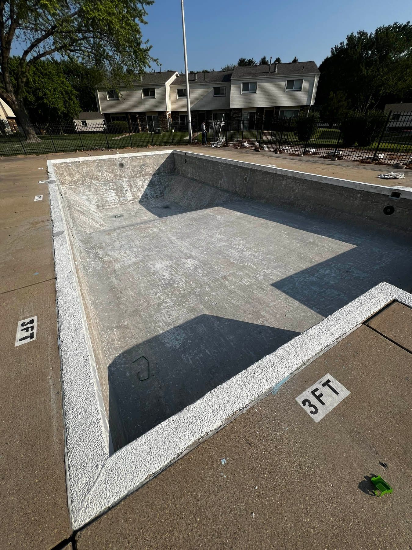 Empty concrete swimming pool with a white trim and depth markers. Apartment buildings visible in the background under a bright blue sky.