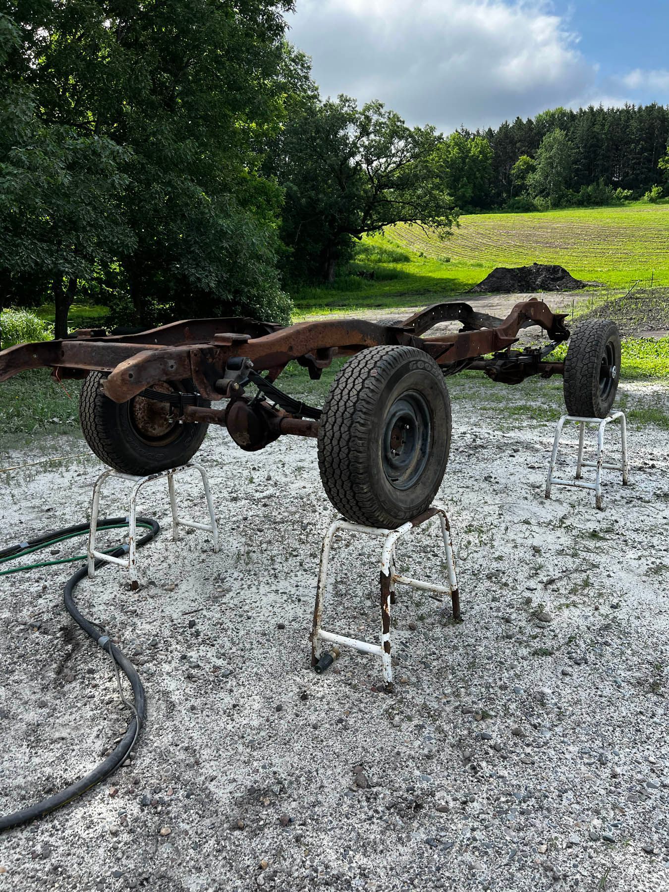 A rusty car chassis with tires rests on supports outdoors, amidst gravel and green foliage.
