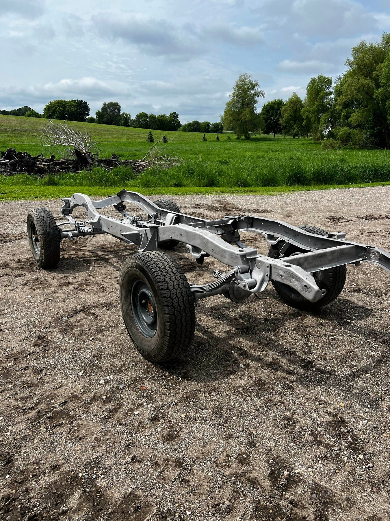 A light gray truck chassis sits on a gravel surface. Green field and cloudy sky are in the background.
