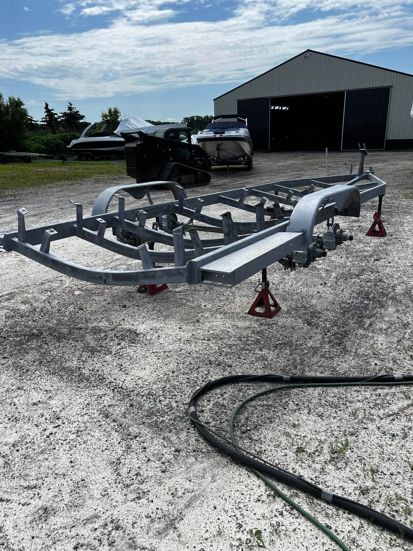A boat trailer is supported by red jacks, with a boat and building visible in the background. Gray trailer in an outdoor setting.