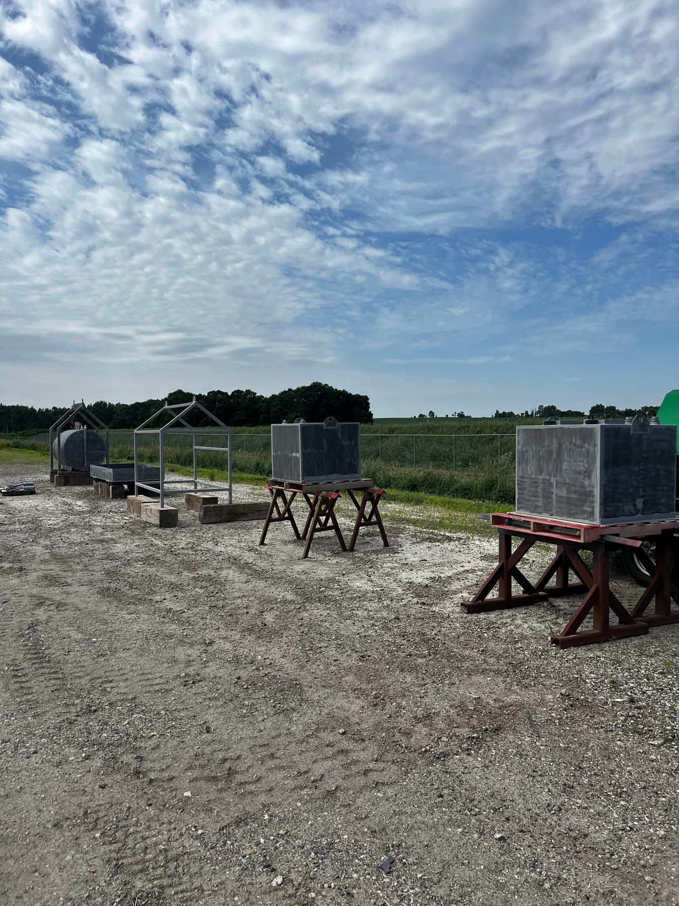 Gravel lot with large stone blocks on sawhorses. A cloudy blue sky and distant field are in the background.