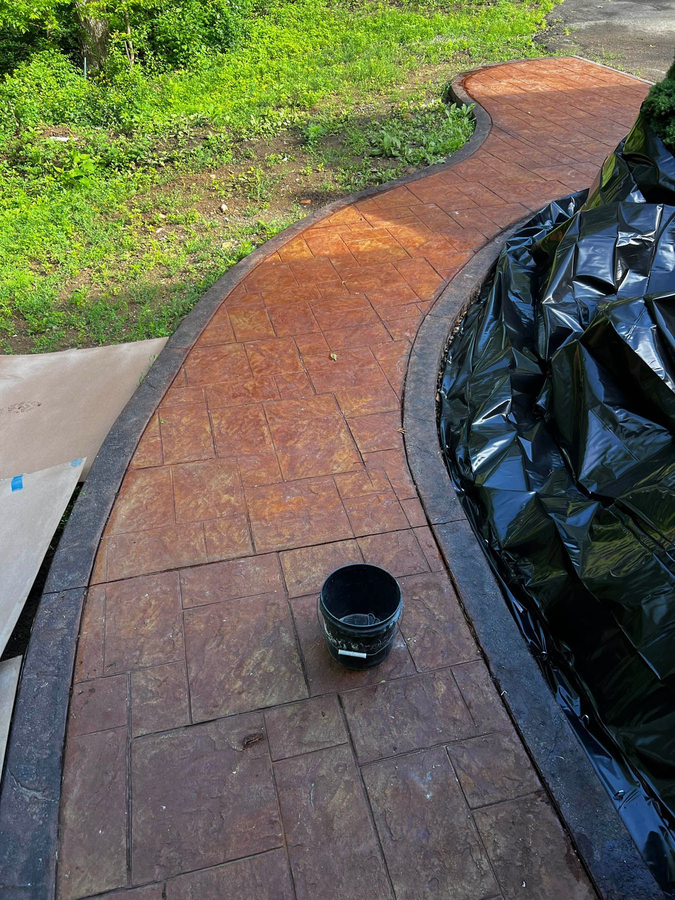 A brown brick-patterned walkway curves along a garden, next to a black-covered structure. A bucket sits on the walkway.
