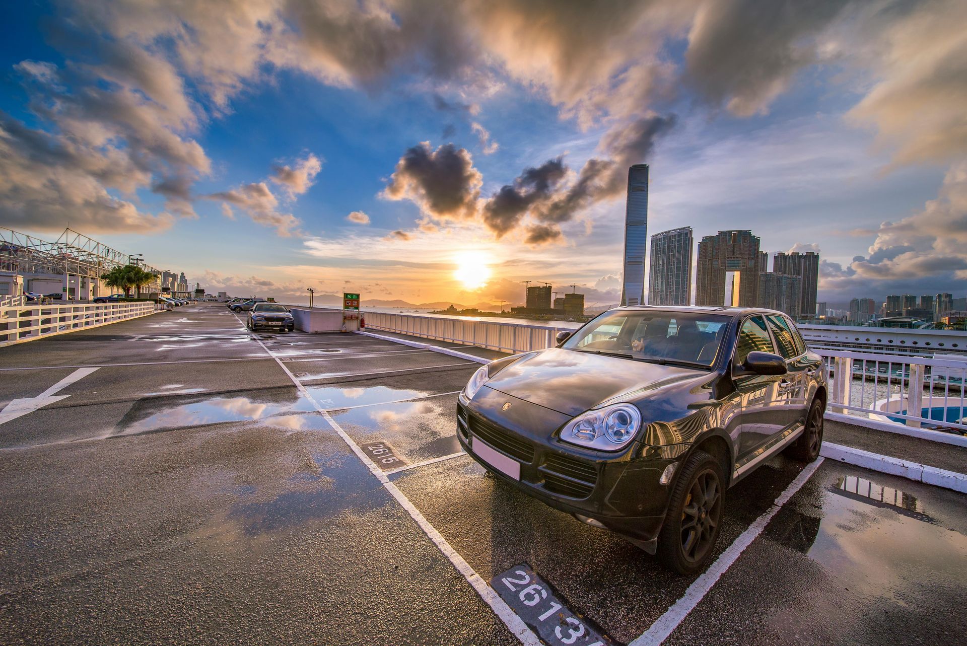 A black Porsche SUV parked in a parking space on a rooftop overlooking a cityscape at sunset with wet pavement.