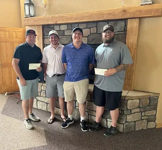 Four men standing in front of a stone fireplace holding checks, likely recipients of a prize.