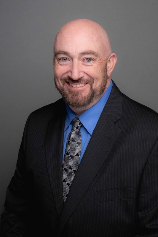 Bald man in a suit smiling. He has a goatee and is wearing a blue shirt and patterned tie.