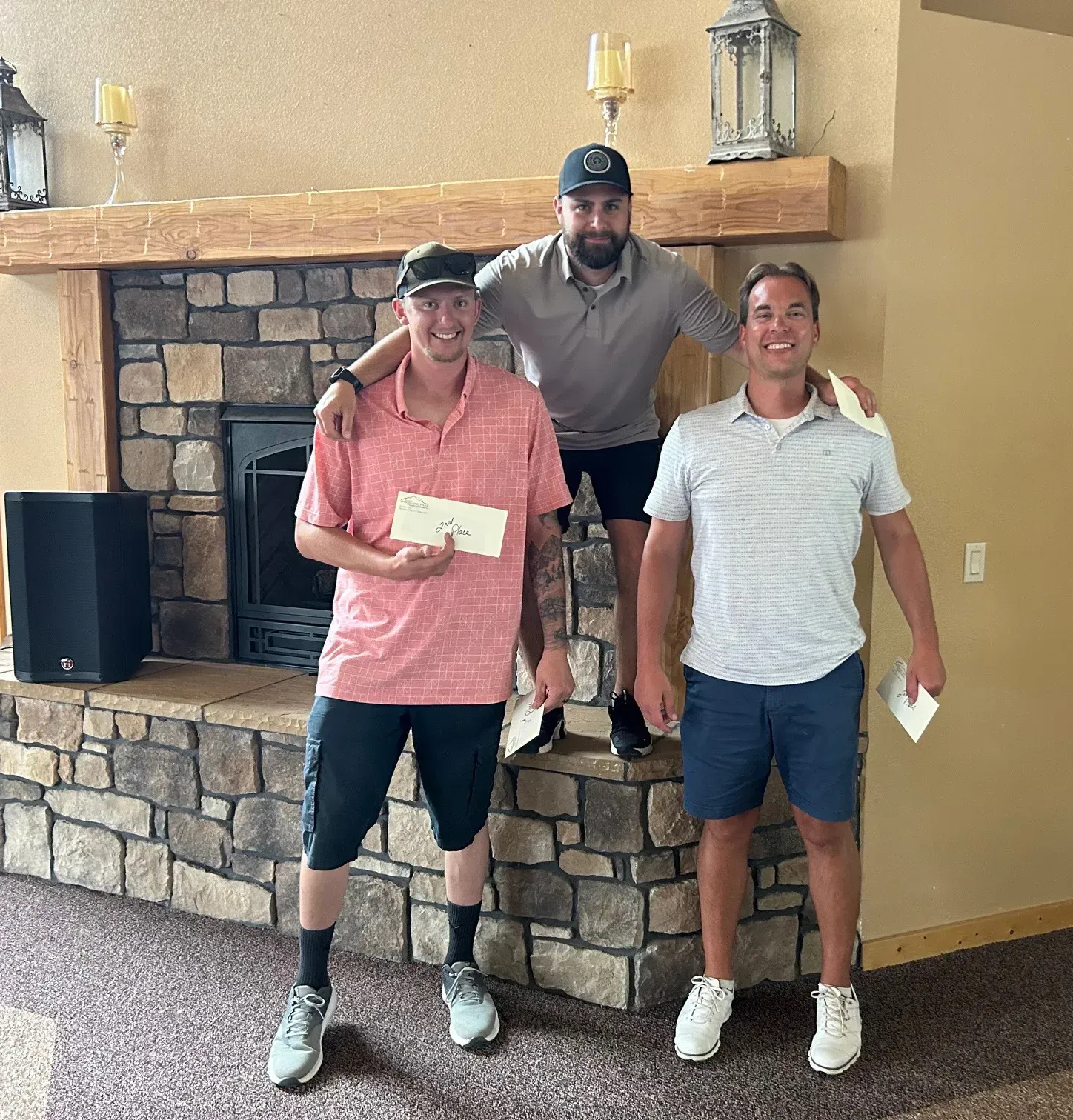 Three men stand near a stone fireplace. One holds a prize, others smile.