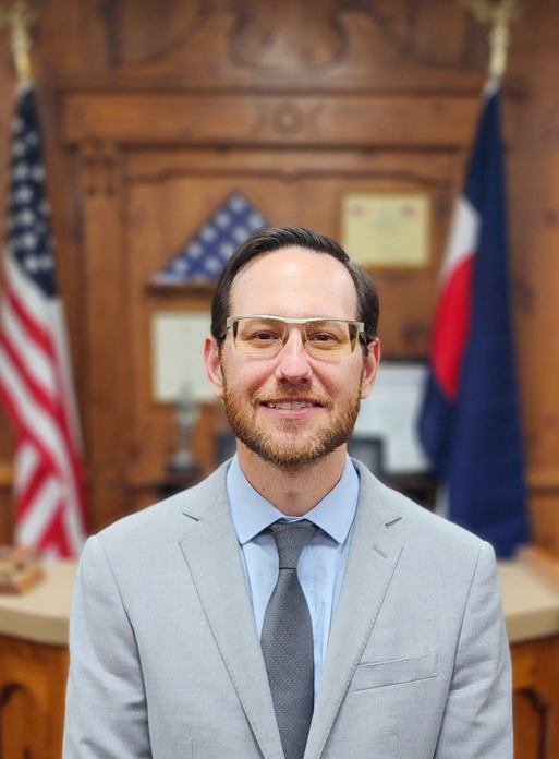 Man in a gray suit and glasses, smiling, with American and Colorado flags behind him.