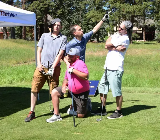 Four men on a golf course. Posing, one wearing pink, one pointing up, with trees and a tent in background.
