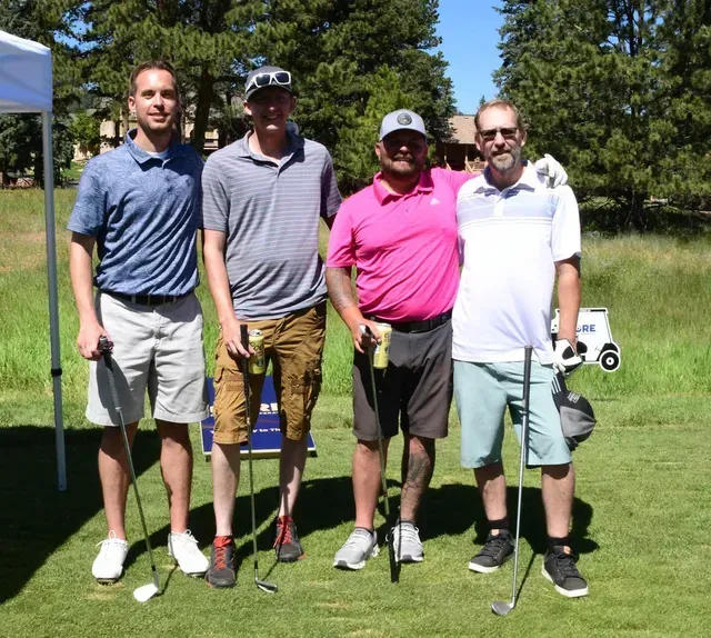 Four men on a golf course, posing. They are wearing casual attire, holding clubs and smiling.