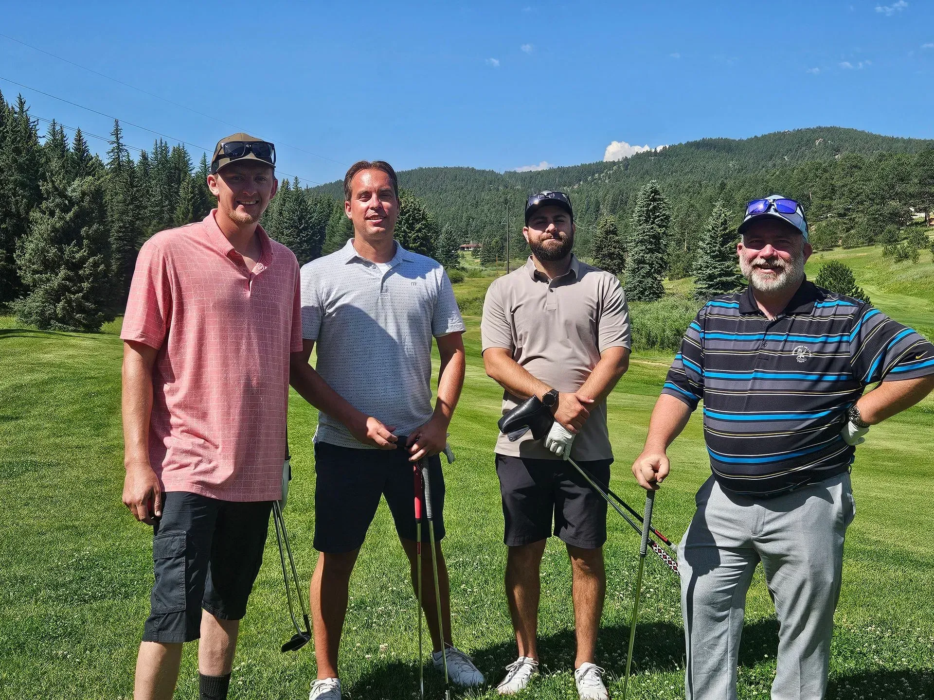 Four men smiling, holding golf clubs, on a sunny golf course with mountains in the background.