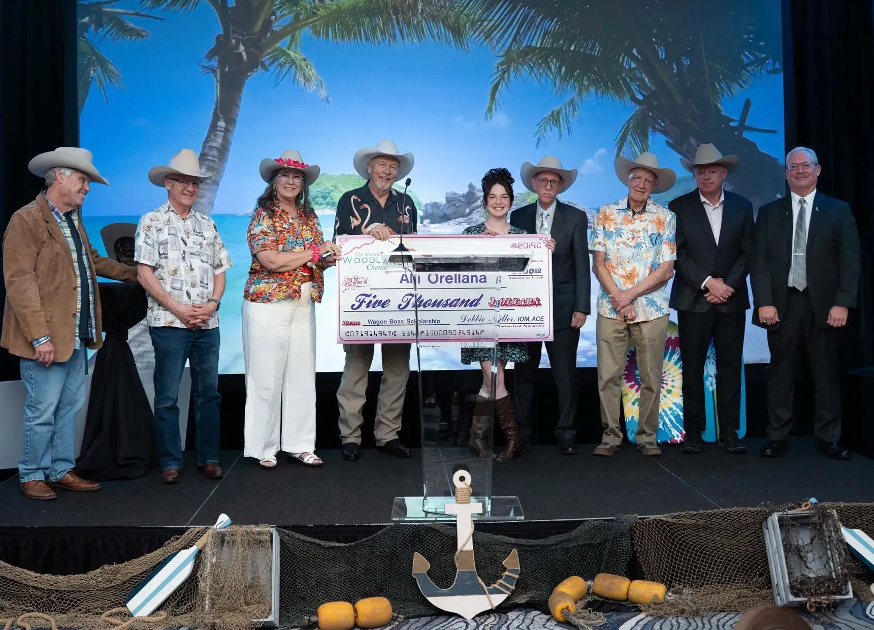 A group of people in cowboy hats hold a large check on a stage with a beach backdrop.