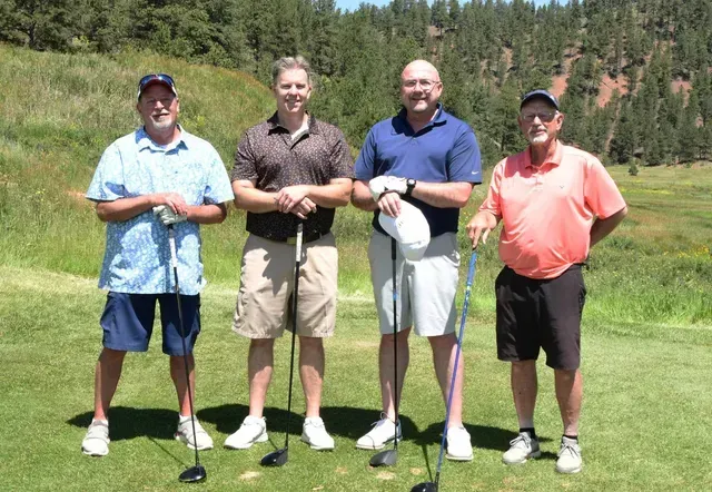 Four men on a golf course, holding clubs. They stand in a row on the green under a sunny sky.
