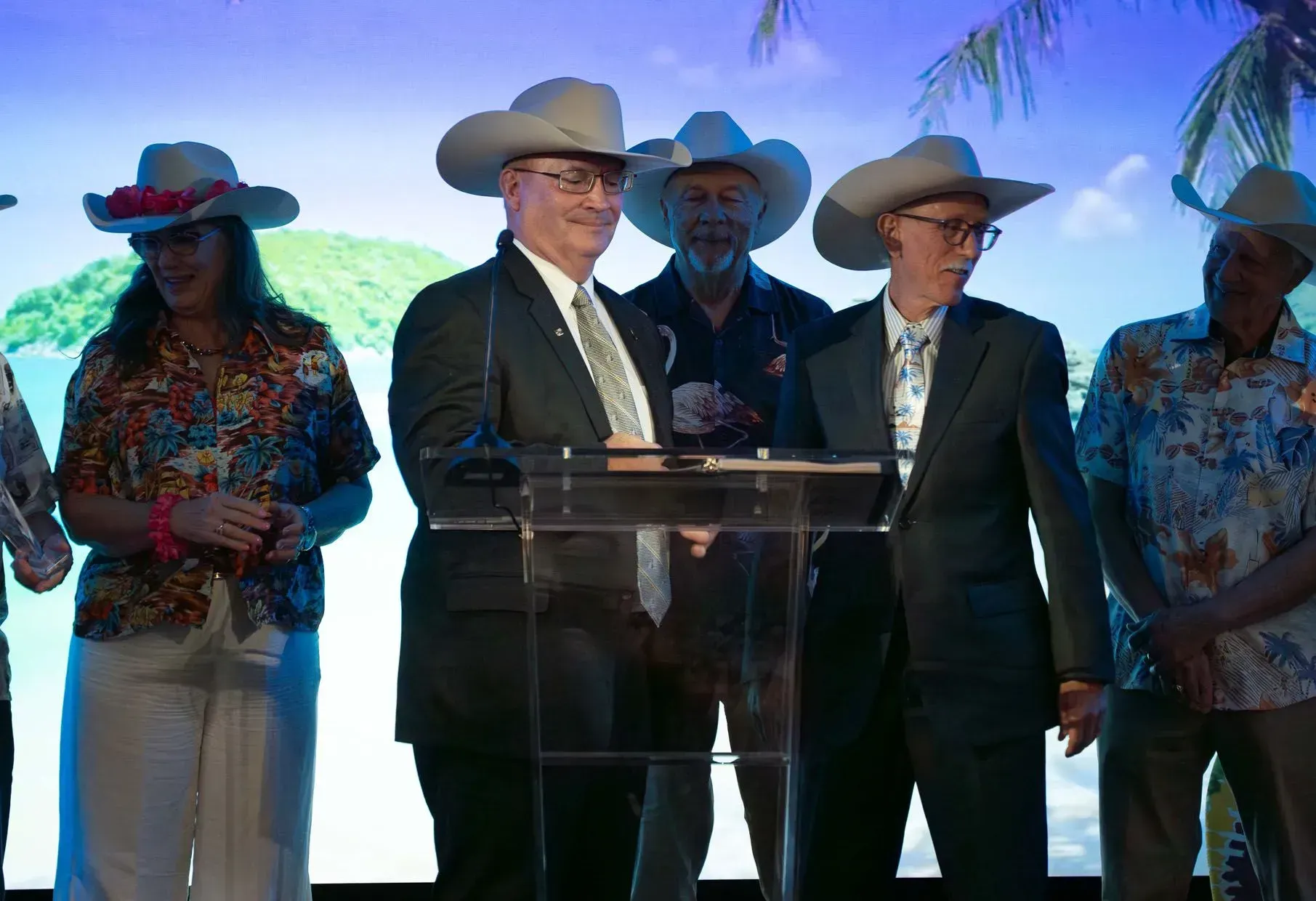 People in cowboy hats at a podium, some in suits and others in Hawaiian shirts, with a tropical backdrop.