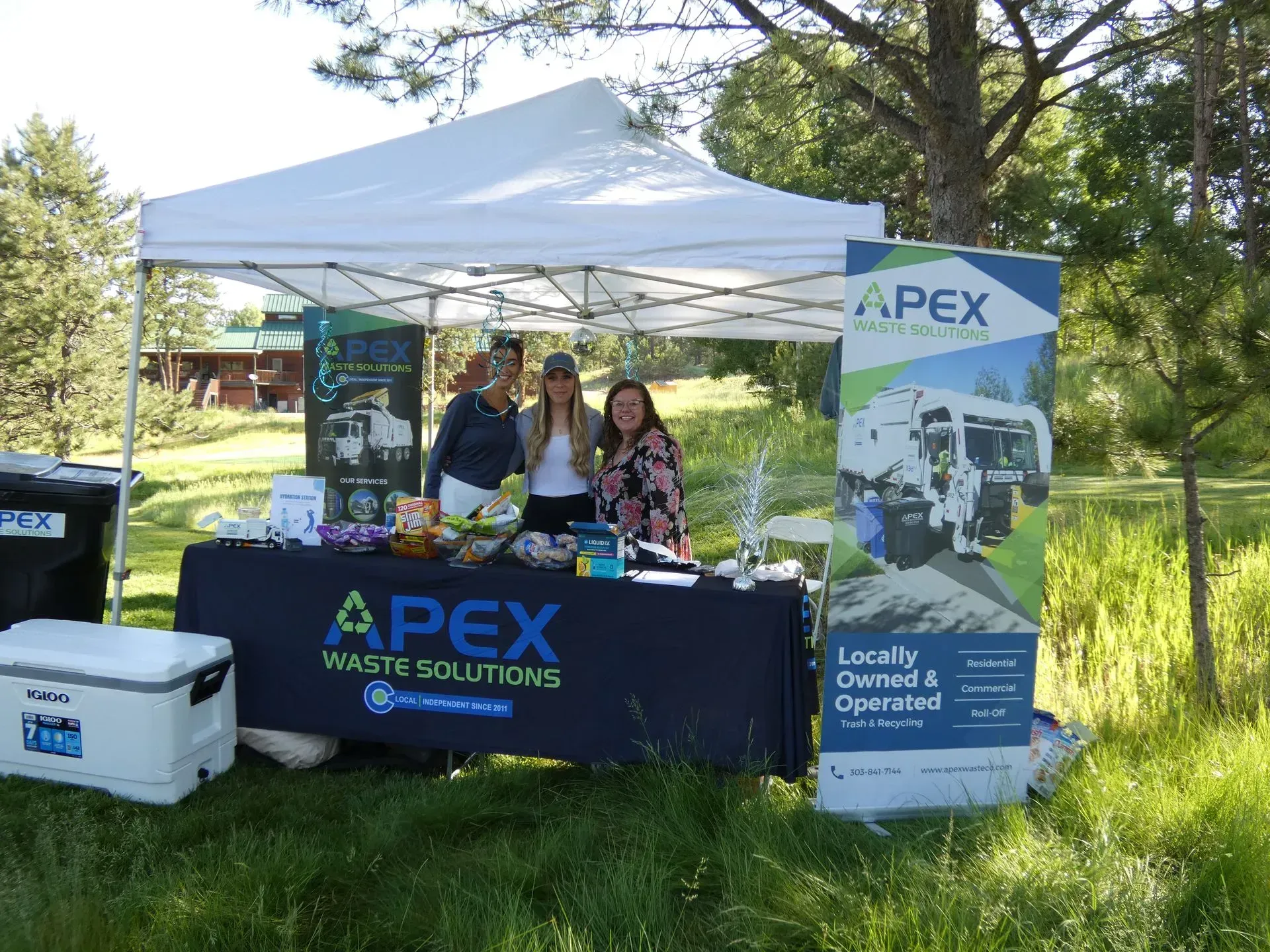 Three women at an outdoor APEX Waste Solutions booth with a banner, snacks, and a cooler.