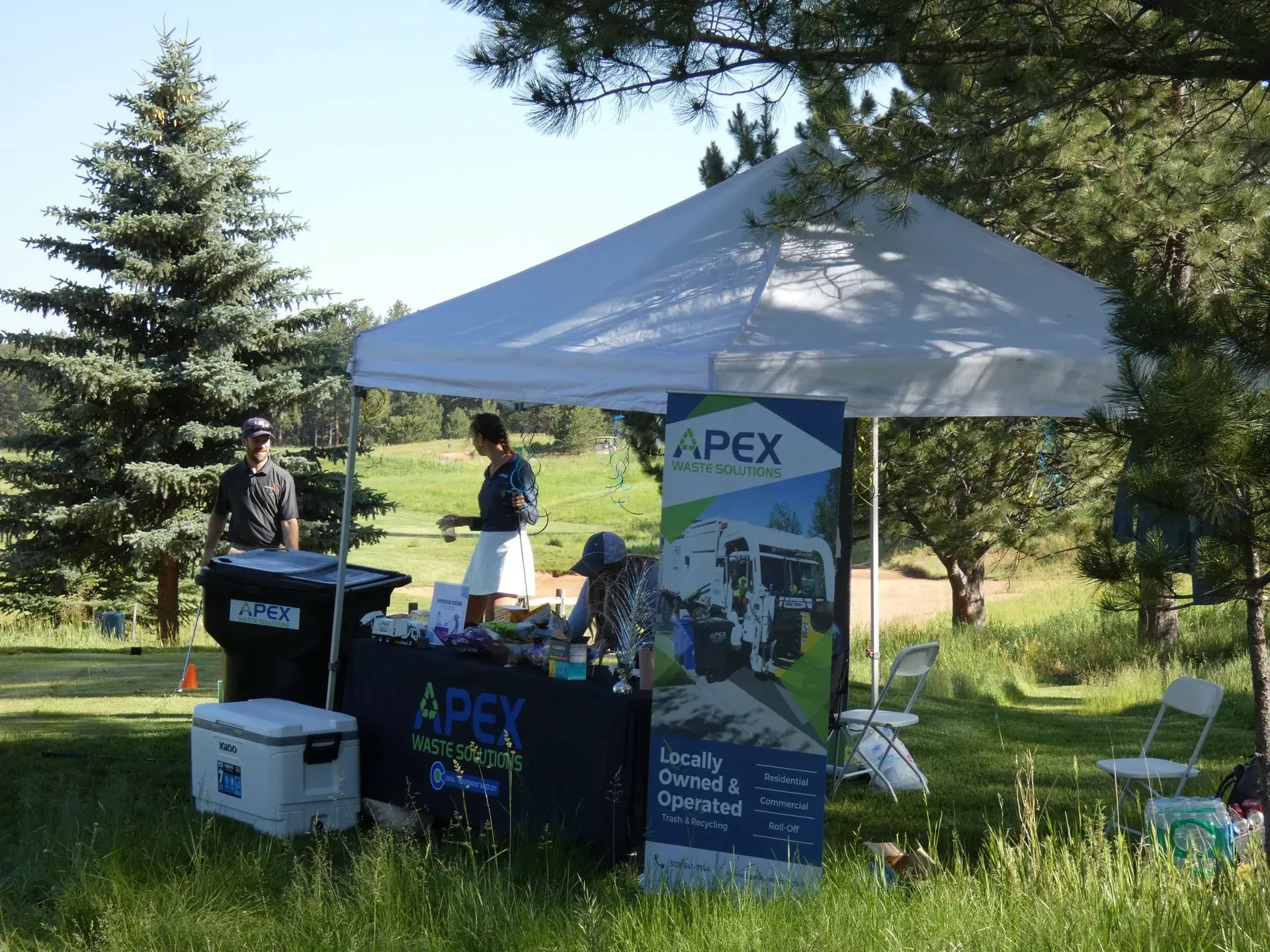 APEX booth on a golf course with three people. Signage and a cooler are visible in a sunny outdoor setting.