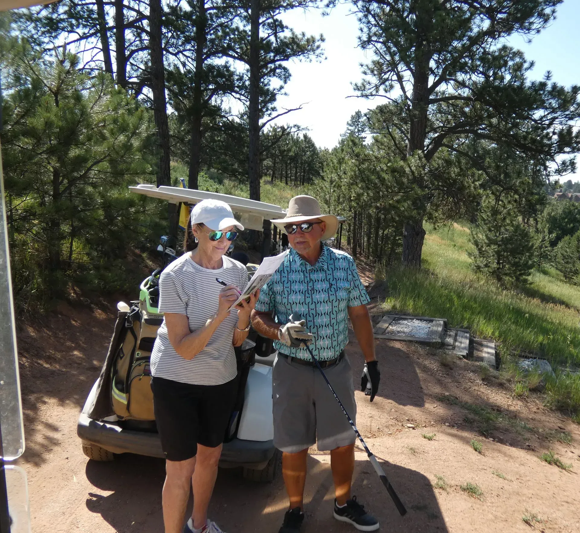 Woman and man at a golf course, looking at a scorecard. Golf cart, trees, and sunny outdoor setting.