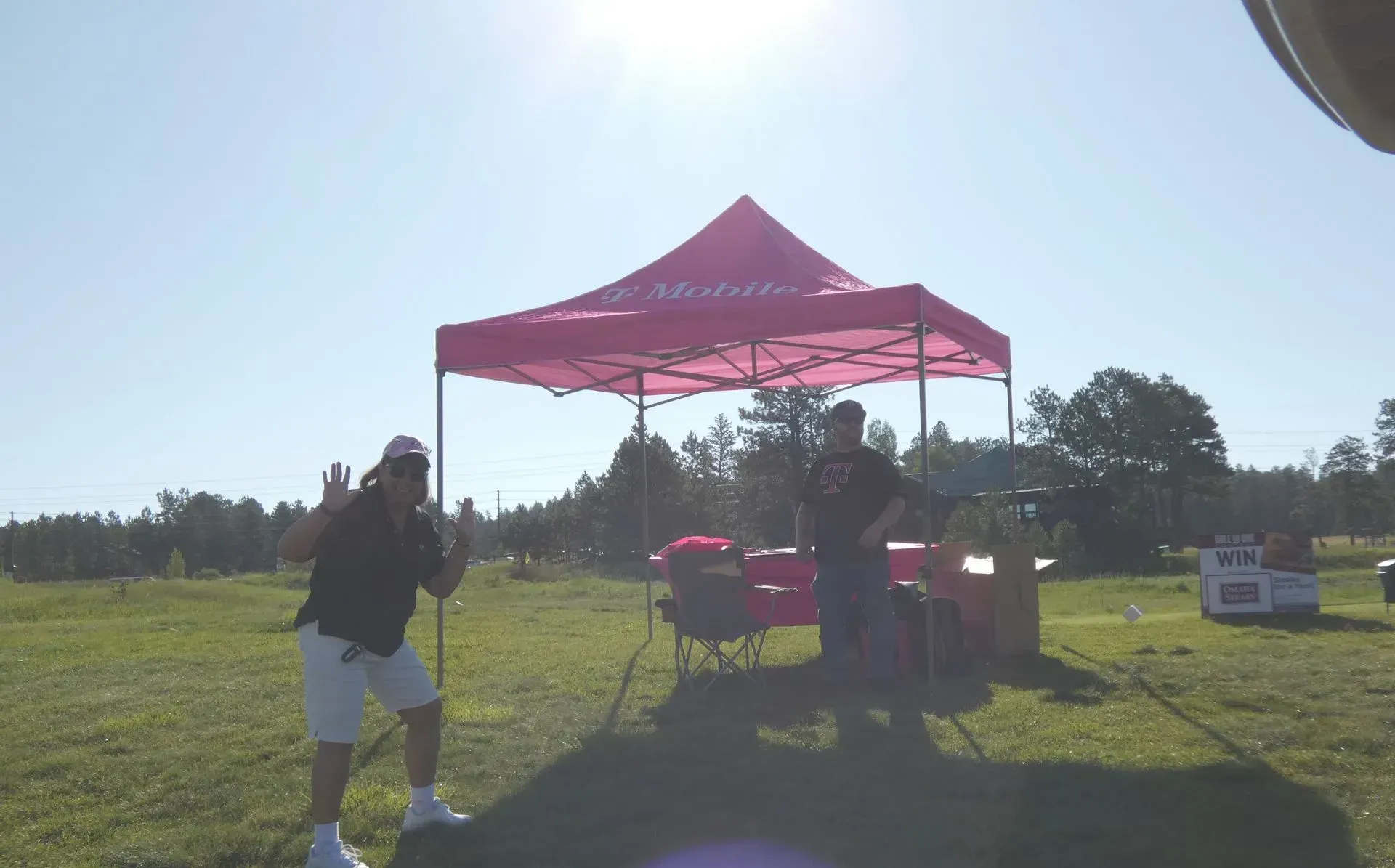 Two people pose under a pink tent on a sunny field. One gestures, the other stands behind a table.