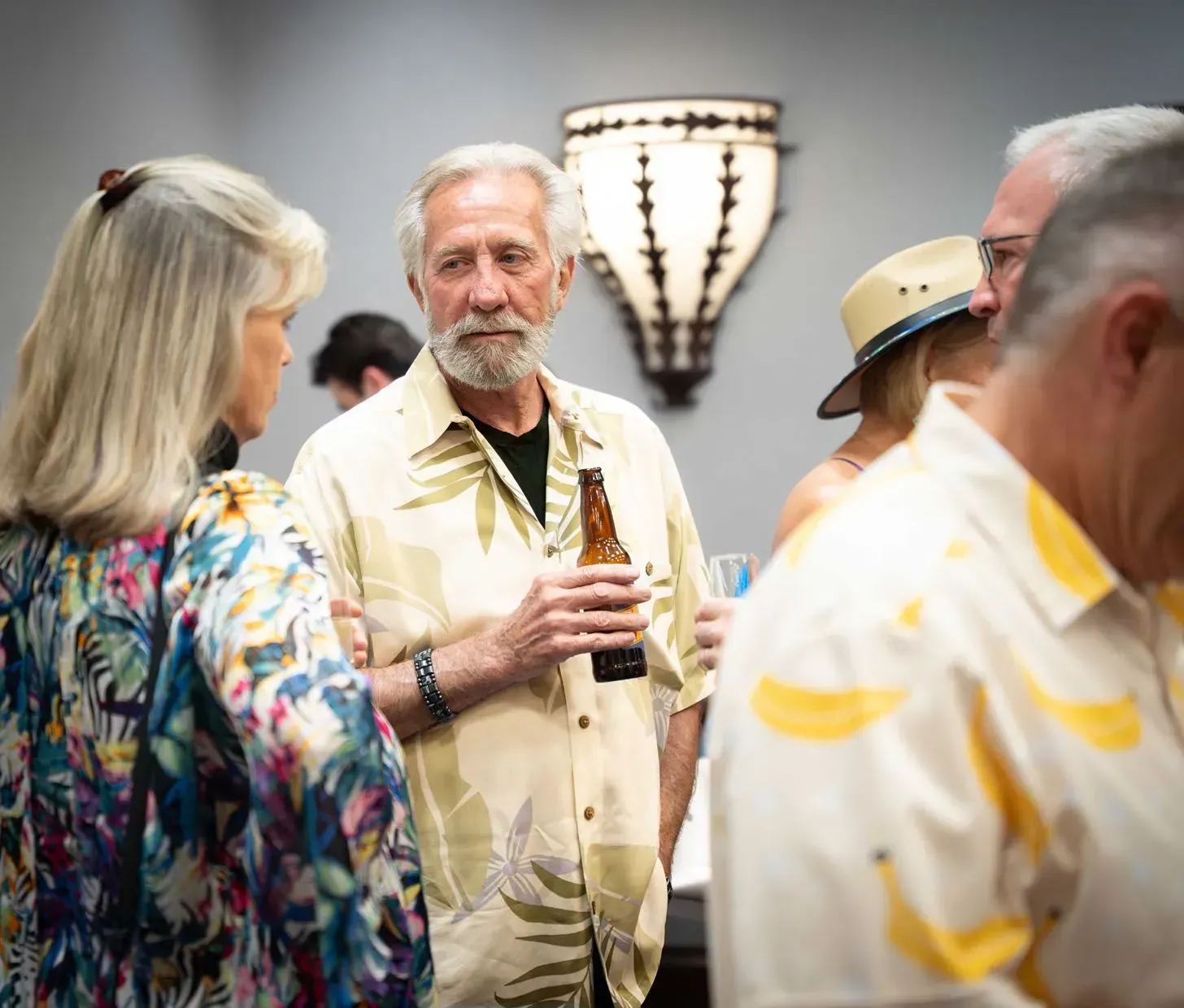 Group of people at a gathering, man holding a beer.  Some wear floral shirts, talking in a room with unique lighting.