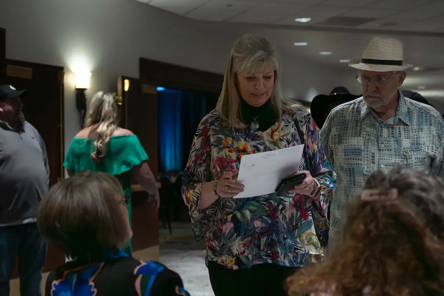 Woman in floral shirt reading to a group; man in a hat looks on; event hallway.