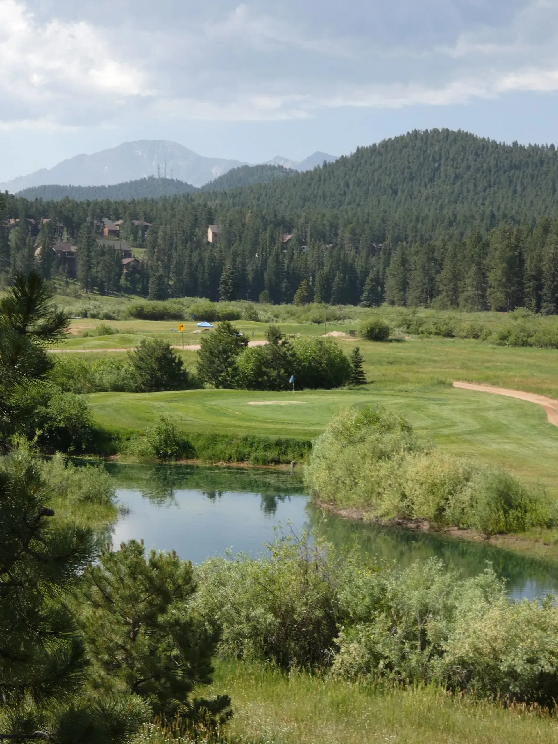 Lush green valley with a lake and golf course, forested hills in the background. Cloudy sky.