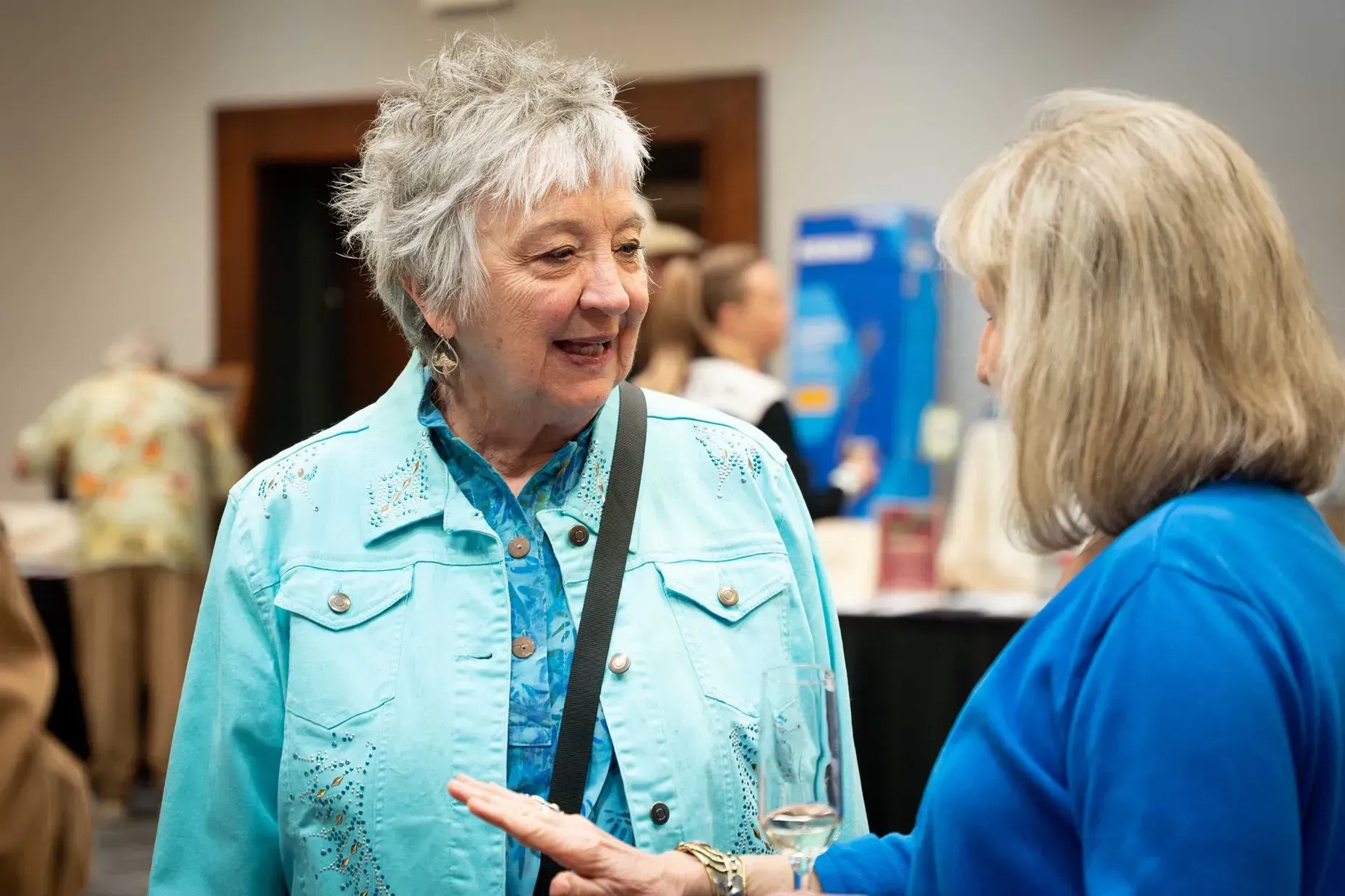 Two women conversing at an event, one wearing a turquoise jacket, the other in blue.